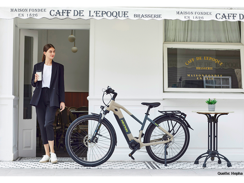 Eine Frau kommt aus einem Cafe zu dem Hepha E-Bike Trekking7.