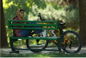 Eine Frau mit Kopftuch sitzt auf einer grünen Parkbank. Vor Ihr ein gelbes Mountainbike.