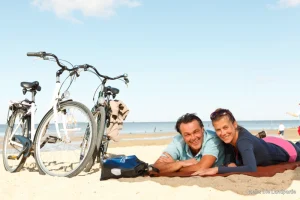 Zwei Fahrräder stehen am Strand, eine Frau und ein Mann liegen auf einer Decke am Strand.